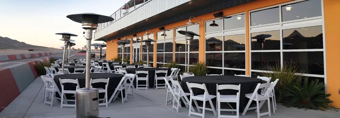 Outdoor dining setup with round tables covered in black cloth and white chairs beside an orange building. Patio heaters and mountains in the background.